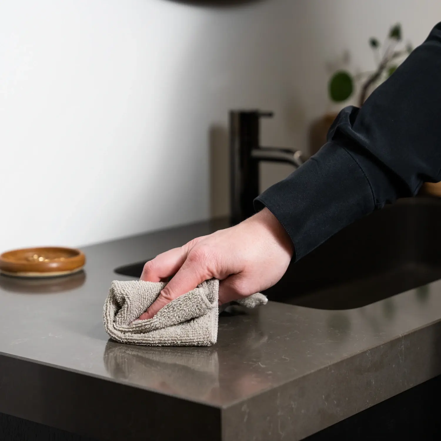 Cleaning and maintenance of bathroom furniture: A person gently wipes a dark bathroom countertop with a beige microfiber cloth to remove dirt and water splashes.