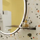 Small bathroom inspiration with a white vanity with black handle and black elegant sink, beautiful round mirror with black edge, offset wall cabinets, and terrazzo wall and floor, as well as yellow walls, provide a bold and modern look.